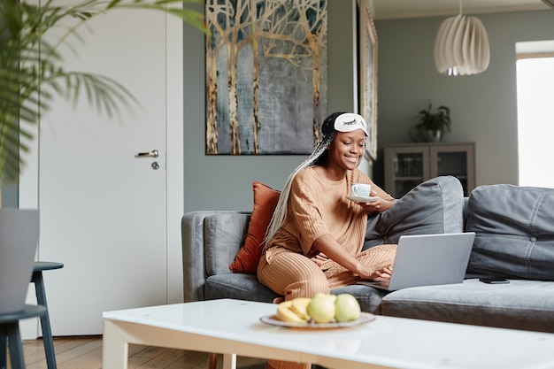 Portret van een jonge Afro-Amerikaanse vrouw die een laptop gebruikt terwijl ze geniet van de ochtend in een gezellig interieur...