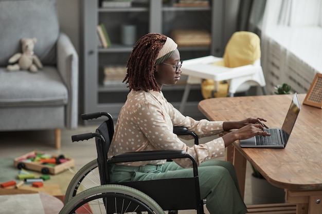 Portret van een jonge Afro-Amerikaanse vrouw die een rolstoel gebruikt terwijl ze thuis werkt met kinderspeelgoed op de achtergrond, kopieer ruimte