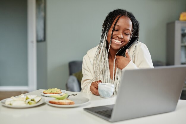 Portret van een jonge Afro-Amerikaanse vrouw die thuis videochat gebruikt terwijl ze 's ochtends geniet van het ontbijt...