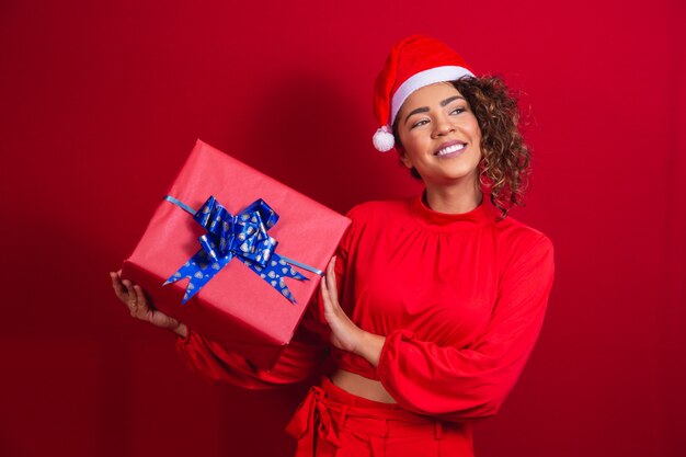 Portret van een jonge afrovrouw die in de hoed van de Kerstman een gift op rode achtergrond houdt. kerstnacht concept