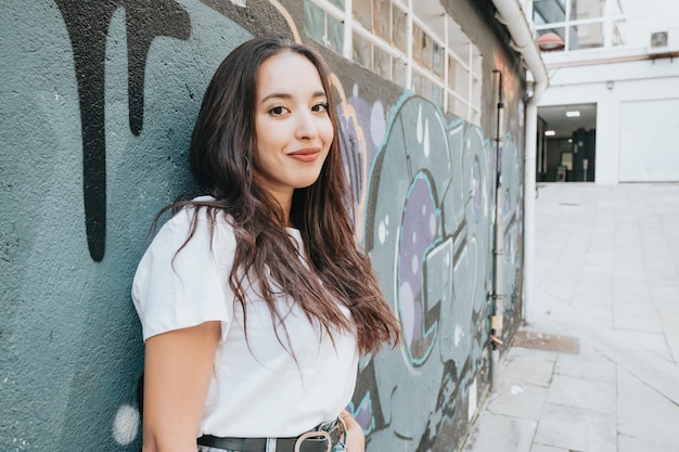 Portret van een jonge Arabische vrouw die een wit overhemd en een blauwe spijkerbroek draagt en naar de camera glimlacht op een bakstenen muur met een achtergrond voor het schilderen van graffiti. Straatlevensstijl, cool trendy. Sociaal netwerk concept