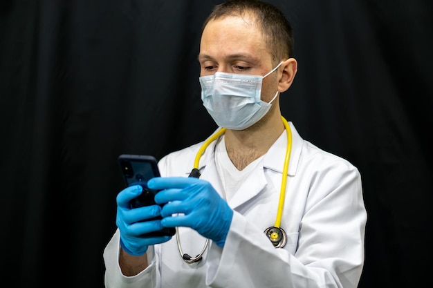 Portret van een jonge arts in een witte jas op een zwarte achtergrond Een arts met een masker en blauwe handschoenen houdt een mobiele telefoon in zijn handen