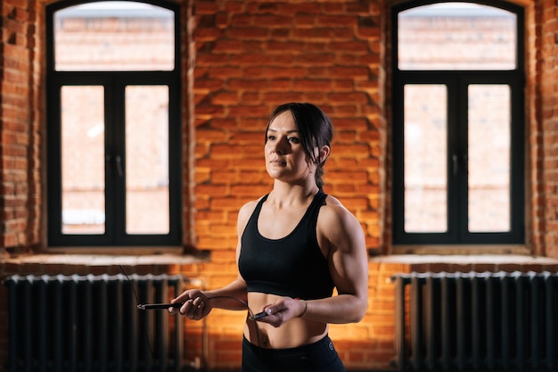 Portret van een jonge, atletische fitnessvrouw met een sterk mooi lichaam in zwarte sportkleding die springtouw vasthoudt terwijl ze een training volgt