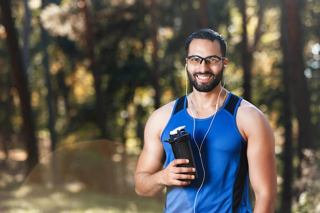 Portret van een jonge atletische multiculturele man met een sportieve bril die pauze heeft en water drinkt in het zonnige park, een gezonde levensstijl en mensenconcept