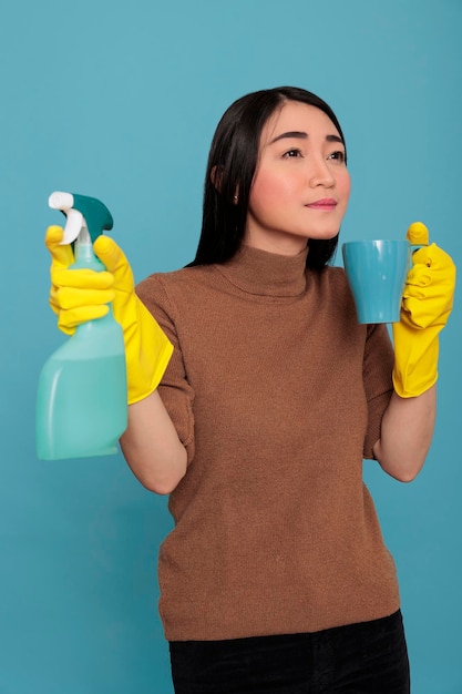 Portret van een jonge aziatische denkende vrouw die koffie drinkt in een gele handschoen terwijl ze aan de andere kant sproeit op een blauwe achtergrond, huisconcept schoonmaken, verfrissing thee tijdens de pauze