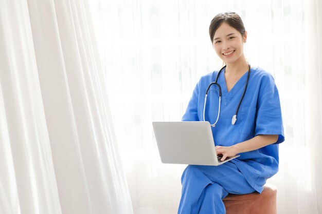 Portret van een jonge Aziatische gelukkige verpleegster met medische scrubs die naar de camera kijkt met een stethoscoop en een laptop gebruikt terwijl ze zit met een wit gordijn op de achtergrond Afbeelding met kopieerruimte