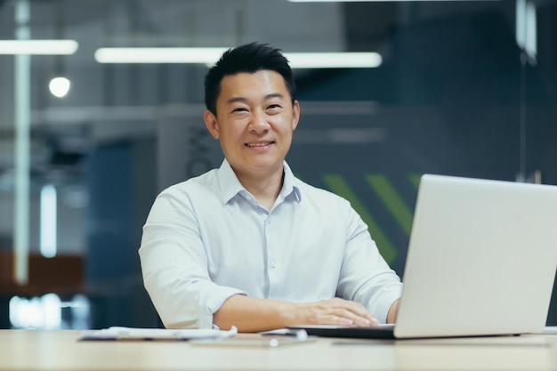 Portret van een jonge aziatische man die glimlachend naar de camera kijkt terwijl hij aan het bureau op kantoor zit