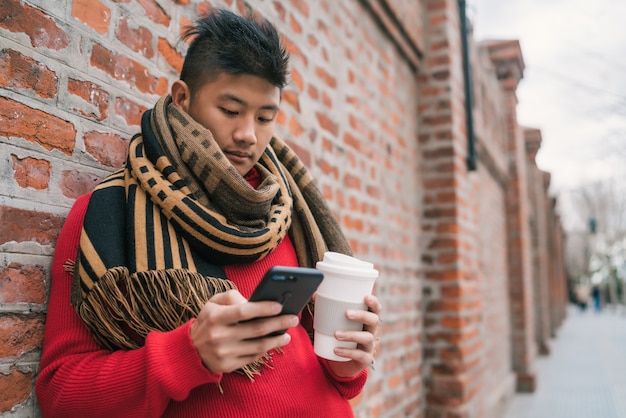 Portret van een jonge Aziatische man die zijn mobiele telefoon gebruikt terwijl hij een kopje koffie vasthoudt, buiten op straat