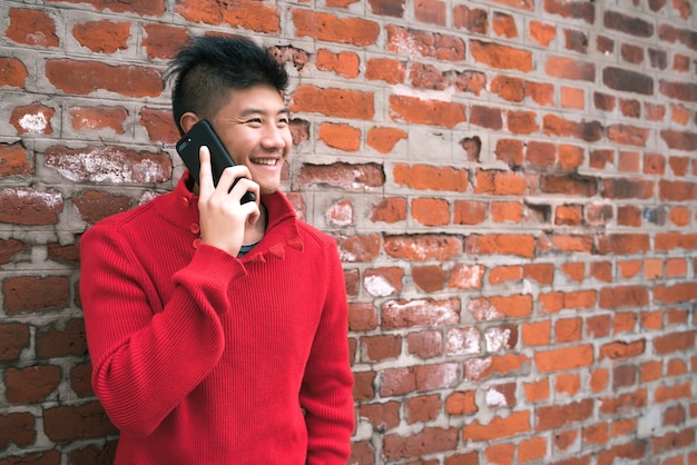 Portret van een jonge Aziatische man praten aan de telefoon buiten tegen bakstenen muur. Communicatie concept.