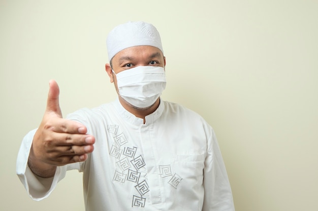 portret van een jonge aziatische moslimman met een masker dat hij een handdruk aanbiedt en glimlacht?