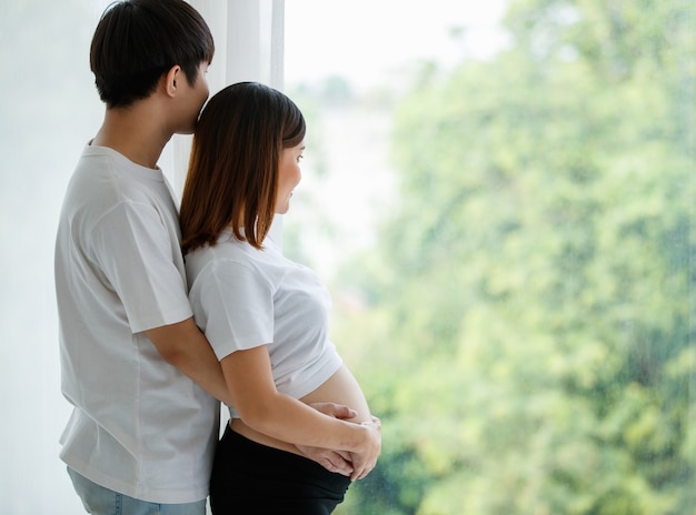 Portret van een jonge Aziatische paar man en zwangere vrouw gekleed terloops glimlachend en samen bij het raam staan met een warm wit licht van buiten. Familieconcept.