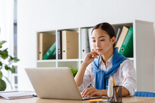 Portret van een jonge aziatische secretaresse die aan tafel zit en een laptop gebruikt terwijl ze op kantoor werkt