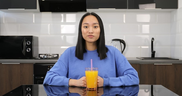 Portret van een jonge Aziatische vrouw die sinaasappelsap drinkt. Een aantrekkelijk multiraciaal brunette meisje zit in de keuken aan tafel. Gezond eten.