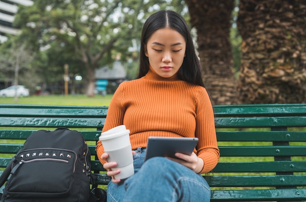 Portret van een jonge Aziatische vrouw met behulp van haar digitale tablet terwijl ze een kopje koffie in het park buiten houdt.