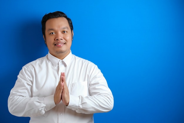 Portret van een jonge Aziatische zakenman met een wit overhemd die naar de camera kijkt, glimlacht en een Aziatisch begroetingsgebaar toont dat op een blauwe achtergrond wordt geïsoleerd