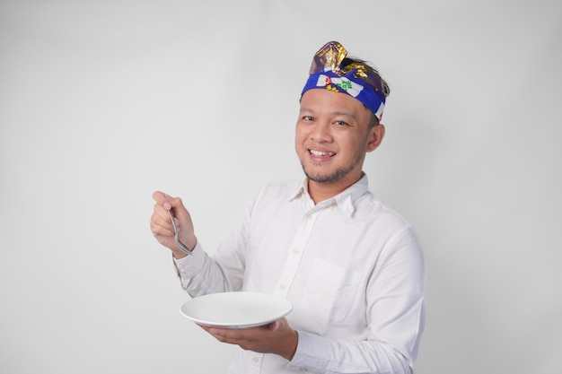 Portret van een jonge Balinese man in wit hemd en traditionele hoofddoek die een leeg bord met kopieerruimte toont en presenteert terwijl hij vrolijk naar de camera glimlacht