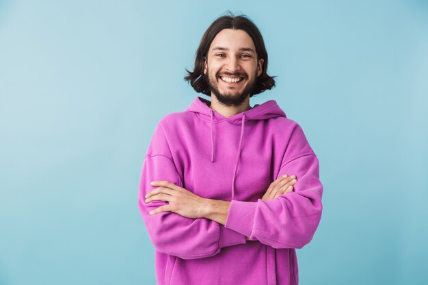 Portret van een jonge, bebaarde brunette man met een hoodie die geïsoleerd over een blauwe muur staat, met gevouwen armen