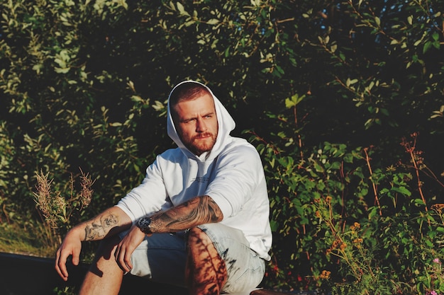 Portret van een jonge, bebaarde man met tatoeages in een witte jas op het platteland of in het park. man in vrijetijdskleding brengt de avond door in de natuur. concept van stijl, wandelen in de frisse lucht en eenheid met de natuur