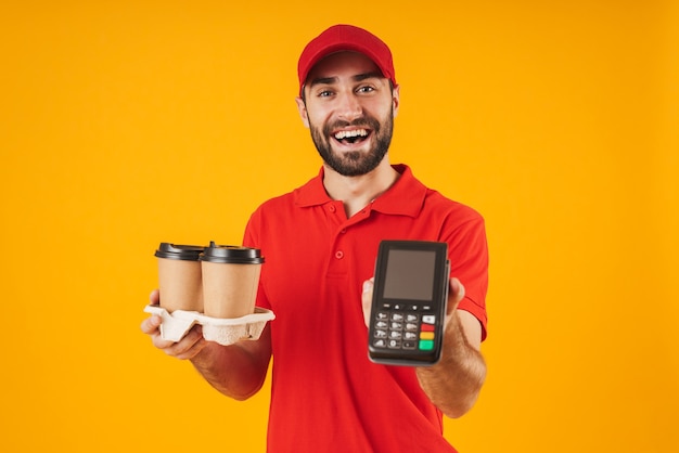 Portret van een jonge bezorger in rood uniform met afhaalkoffiekopjes en betaalterminal geïsoleerd over geel