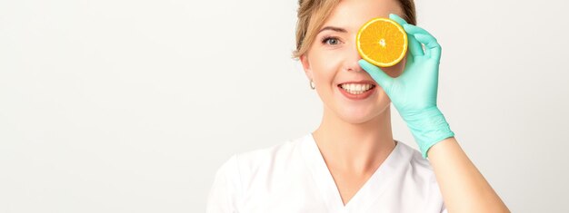 Portret van een jonge blanke glimlachende vrouwelijke schoonheidsspecialiste die het oog bedekt met een sinaasappelschijfje met handschoenen op een witte achtergrond