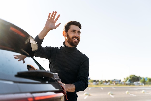 Portret van een jonge blanke man die zijn auto met een microvezel buiten schoonmaakt bij zonsondergang terwijl hij naar iemand op straat zwaait