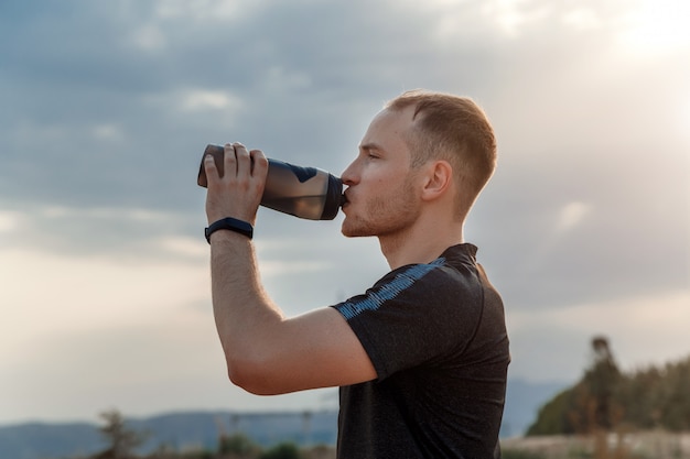 Portret van een jonge blanke man in een zwart t-shirt en zwarte korte broek drinkwater uit een fles