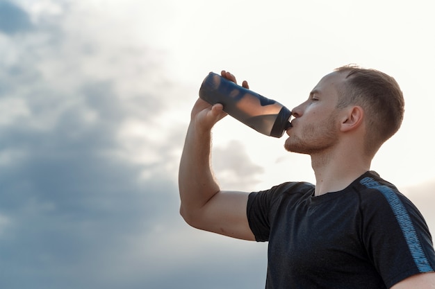 Portret van een jonge blanke man in een zwart t-shirt en zwarte korte broek drinkwater uit een fles