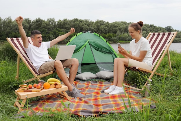 Portret van een jonge blanke man met laptop in de buurt van tent zittend in een opklapbare armstoel met notitieboekje en opgeheven armen schreeuwend en juichend zijn succes afwerkend werk vrouw met behulp van mobiele telefoon