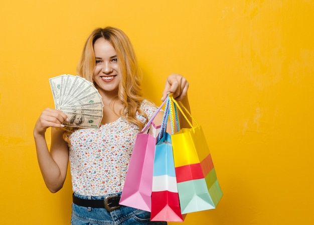 Portret van een jonge blonde gelukkige bankbiljetten van de meisjesholding en het winkelen zak over gele muur