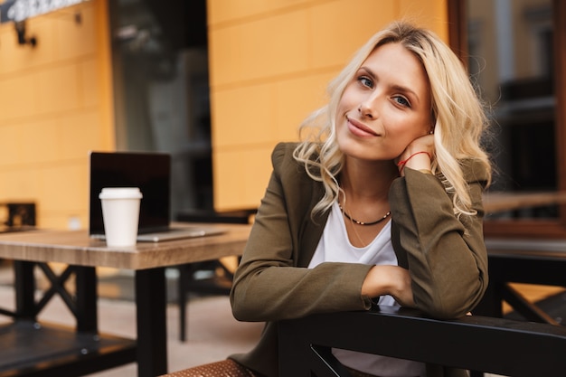 Portret van een jonge blonde vrouw die lacht, zittend in café buiten met afhaalmaaltijden koffie en zilveren laptop