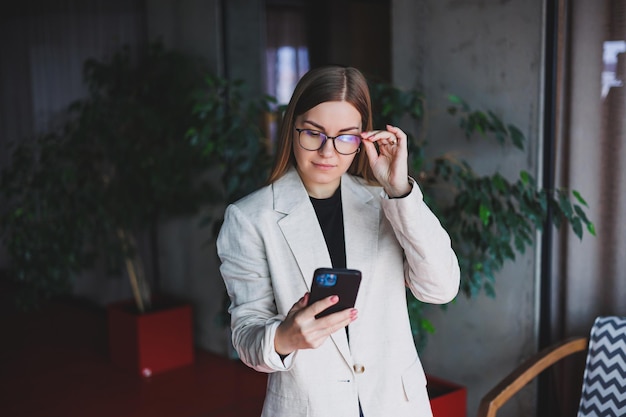 Portret van een jonge blonde vrouw in een beige jas en een bril op haar gezicht een mod-telefoon in de handen van een vrouw Mooi modern blogger-meisje