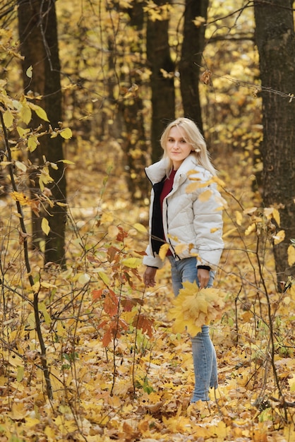 Portret van een jonge blonde vrouw in het herfstbos, met een boeket gele bladeren in haar handen.