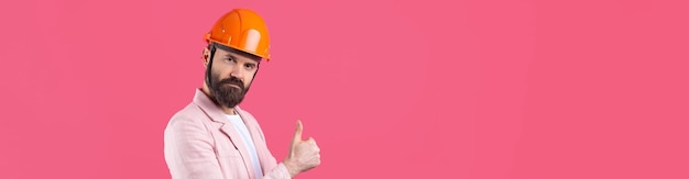Portret van een jonge bouwingenieur die een oranje helm draagt in een roze jas die op een rode studioachtergrond staat. De man wijst met zijn hand