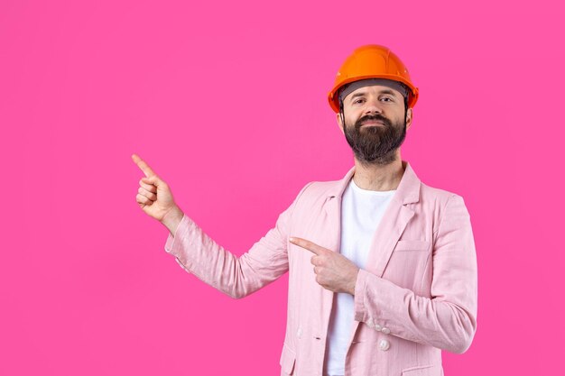 Portret van een jonge bouwingenieur die een oranje helm draagt in een roze jas die op een rode studioachtergrond staat. De man wijst met zijn hand