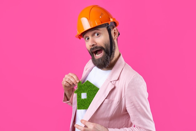 Portret van een jonge bouwingenieur die een oranje helm draagt in een roze jas die op een rode studioachtergrond staat Een man heeft een groen eco-huis