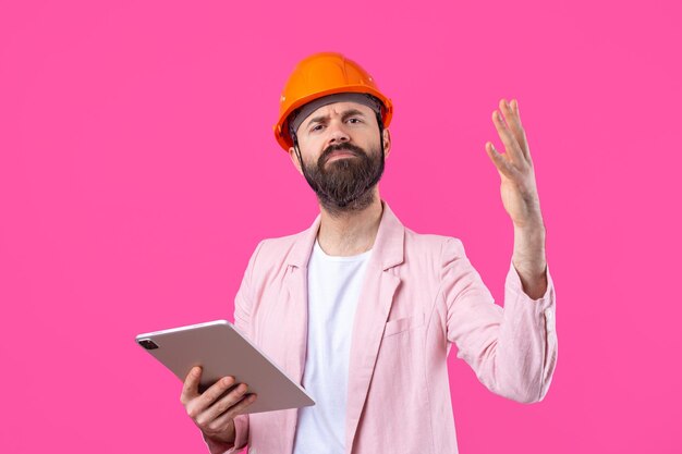 Portret van een jonge bouwingenieur met een oranje helm in een roze jas op een rode studioachtergrond Een man met een tablet in zijn handen