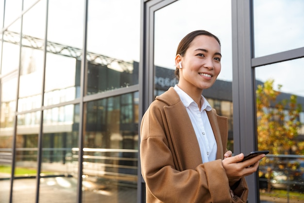 Portret van een jonge brunette aziatische vrouw die een jas draagt met een mobiele telefoon en oordopjes terwijl ze door de stadsstraat loopt