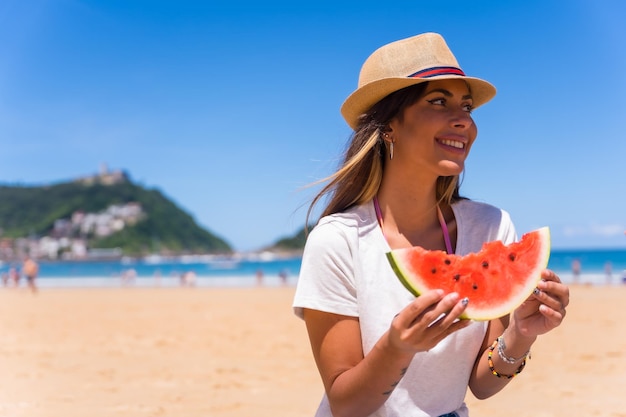 Portret van een jonge brunette blanke vrouw die een watermeloen eet op het strand op zomervakantie met een hoed