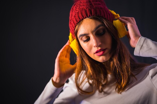 Portret van een jonge brunette Kaukasisch met gele koptelefoon, wit t-shirt en wollen hoed op een zwarte achtergrond, mooi meisje dat naar muziek luistert