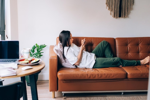 Portret van een jonge brunette vrouw die sms't op haar smartphone terwijl ze thuis op de bank zit.