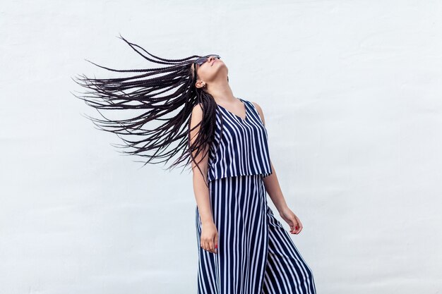 Portret van een jonge brunette vrouw met zwarte afro-amerikaanse dreadlocks kapsel in gestreepte jurk en zonnebril permanent en snel vliegende haar haar dreadlocks. indoor studio opname op witte muur.