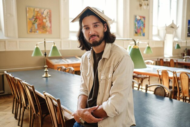 Portret van een jonge casual mannelijke student die bedachtzaam in de camera kijkt terwijl hij aan het dollen is met een boek op het hoofd in de bibliotheek van de universiteit