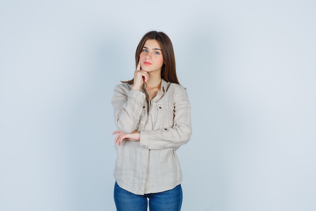 Portret van een jonge dame die de vinger op de wang houdt in casual, jeans en zelfverzekerd vooraanzicht