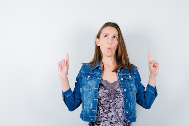 Portret van een jonge dame die omhoog wijst terwijl ze omhoog kijkt in blouse, spijkerjasje en perplex vooraanzicht kijkt