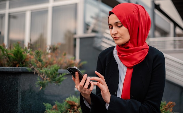 Portret van een jonge, gelukkige moslim zakenvrouw in hijab met behulp van een mobiele telefoon in de buurt van het kantoorgebouw