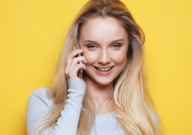 Portret van een jonge gelukkige vrouw die aan de telefoon praat en naar de camera kijkt over gele achtergrond