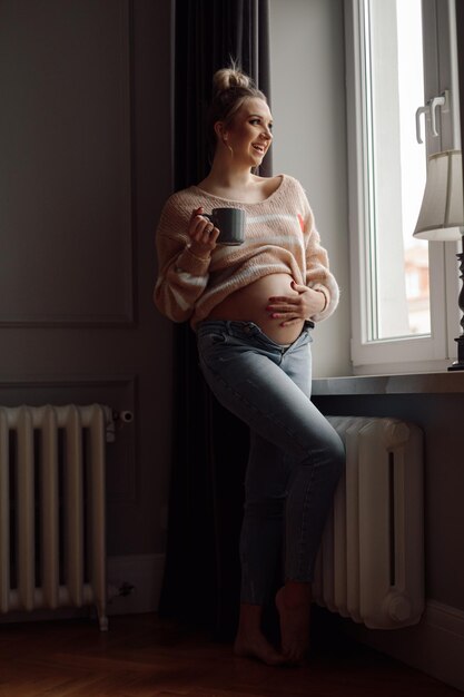 Portret van een jonge, gelukkige zwangere vrouw die bij het raam staat en een beker streelt die haar naakte buik aanraakt
