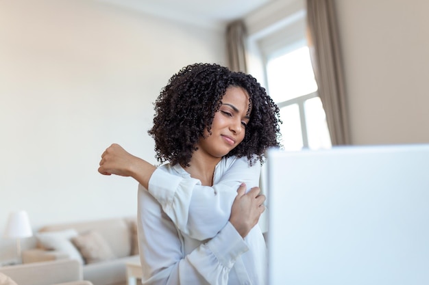 Portret van een jonge gestresste vrouw die aan een thuisbureau voor een laptop zit en een pijnlijke rug aanraakt met een gepijnigde uitdrukking die lijdt aan rugpijn na het werken op de pc