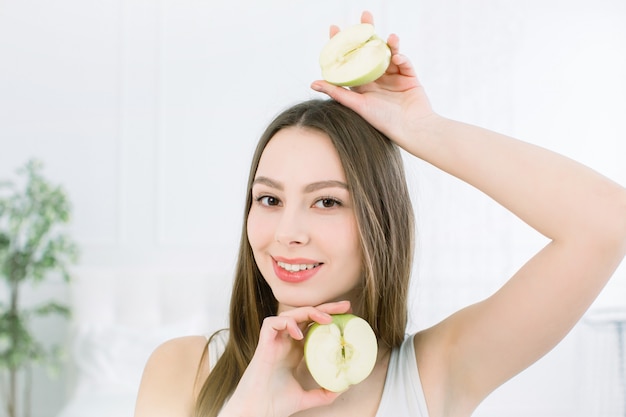 Portret van een jonge glimlachend mooi meisje met twee helften van appels in haar handen, boven en onder het gezicht. vrij kaukasisch meisje dat de helft van appel houdt en op de lichte achtergrond stelt