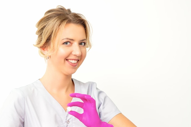 Portret van een jonge glimlachende blanke schoonheidsspecialiste met roze handschoenen die suikerklontjes vasthoudt en naar de camera kijkt tegen een witte achtergrond